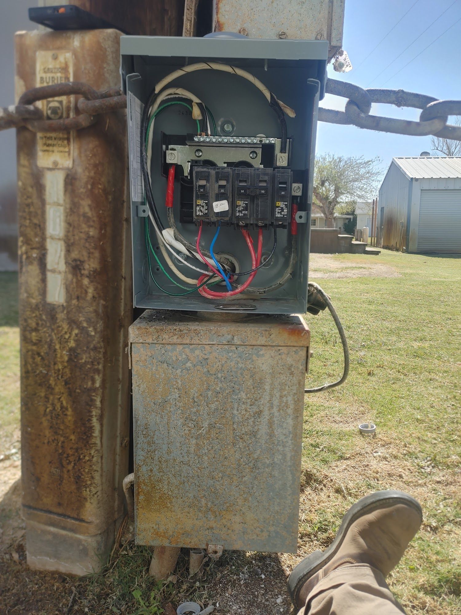 An open electrical panel on a utility pole. Wires and breakers visible.  Rust, grass, and a building are in the background.