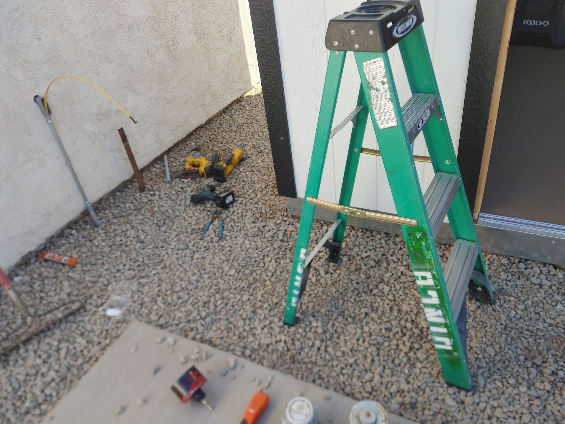 Green ladder and tools on gravel next to a white wall and dark door.
