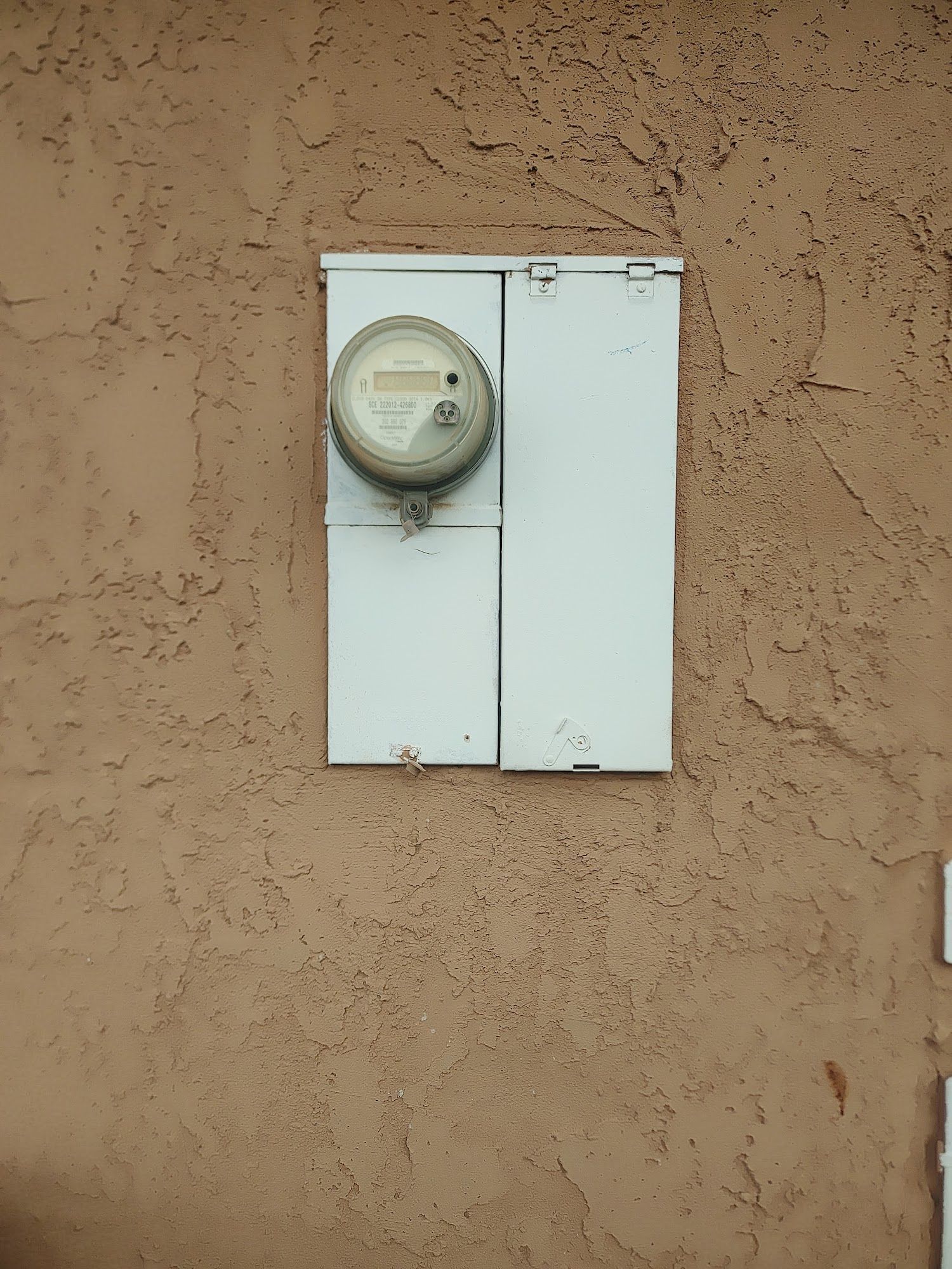 Electric meter in a white metal box, mounted on a textured, tan exterior wall.