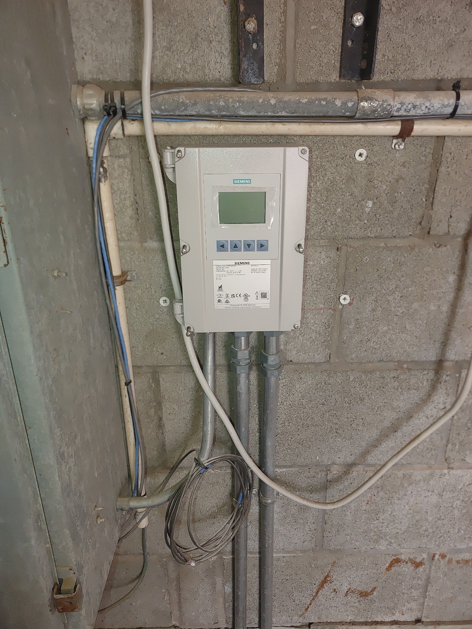 A grey Siemens control panel box with a screen and buttons mounted on a cinder block wall, with wiring.