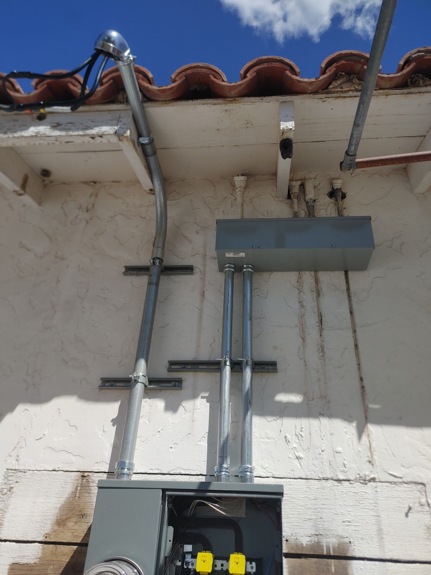 Electrical panel with conduits mounted on a stucco wall beneath a terracotta tile roof.