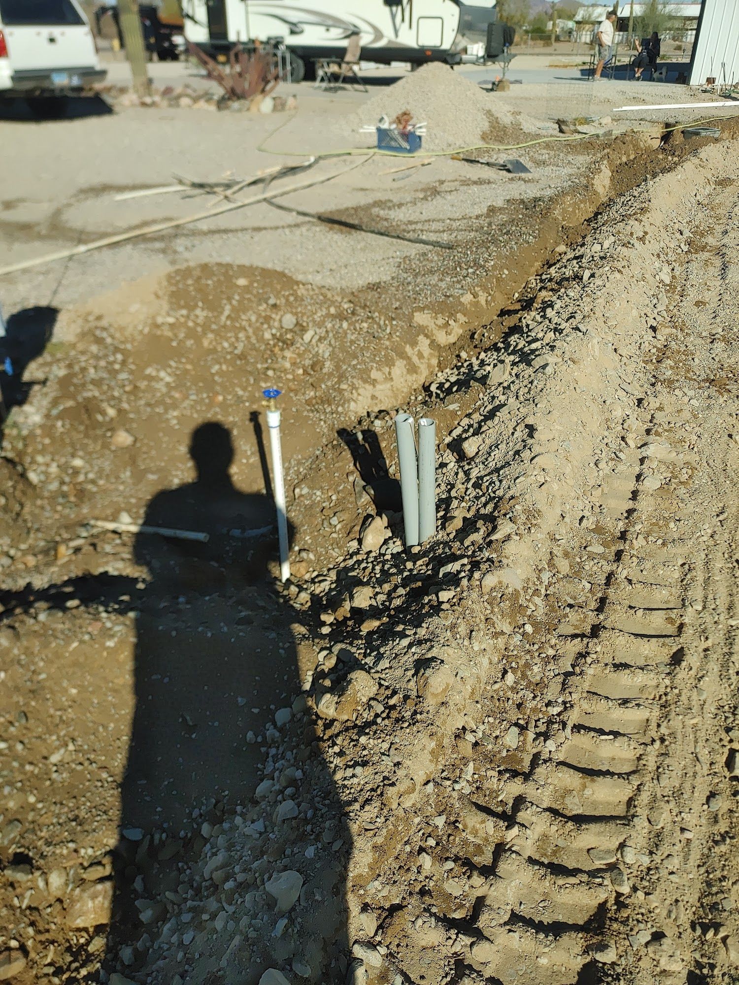 Trench dug next to a road, with PVC pipes and shadow of person holding tool.