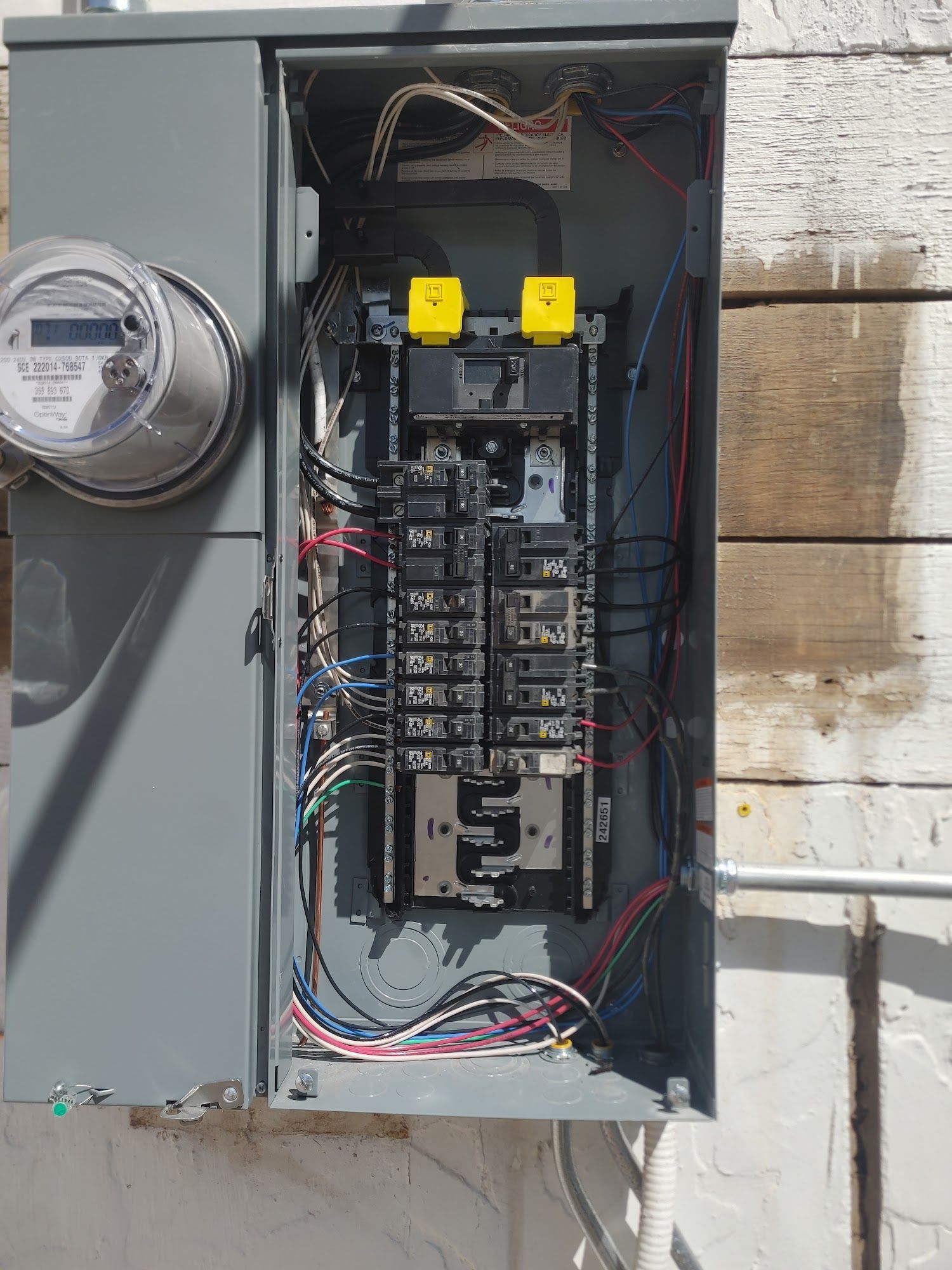 Open electrical panel with circuit breakers and wiring. Gray metal box mounted on a white wall.