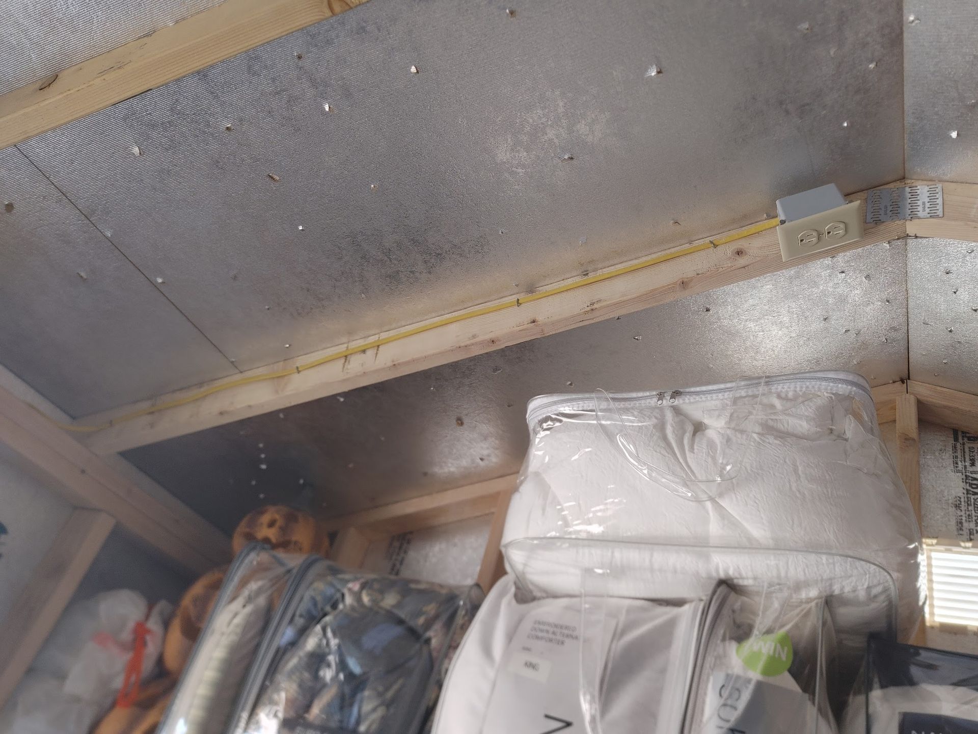 Interior of a shed. Insulation and electrical outlet are visible. Stored items partially obscure view.