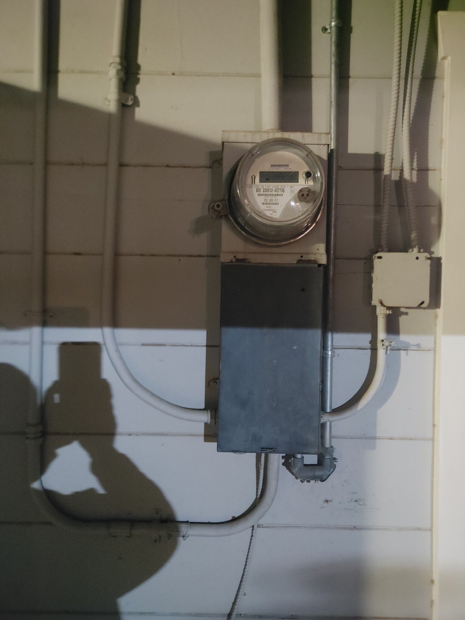 Electrical meter box mounted on a white wall with conduit. Shadow of a person visible.