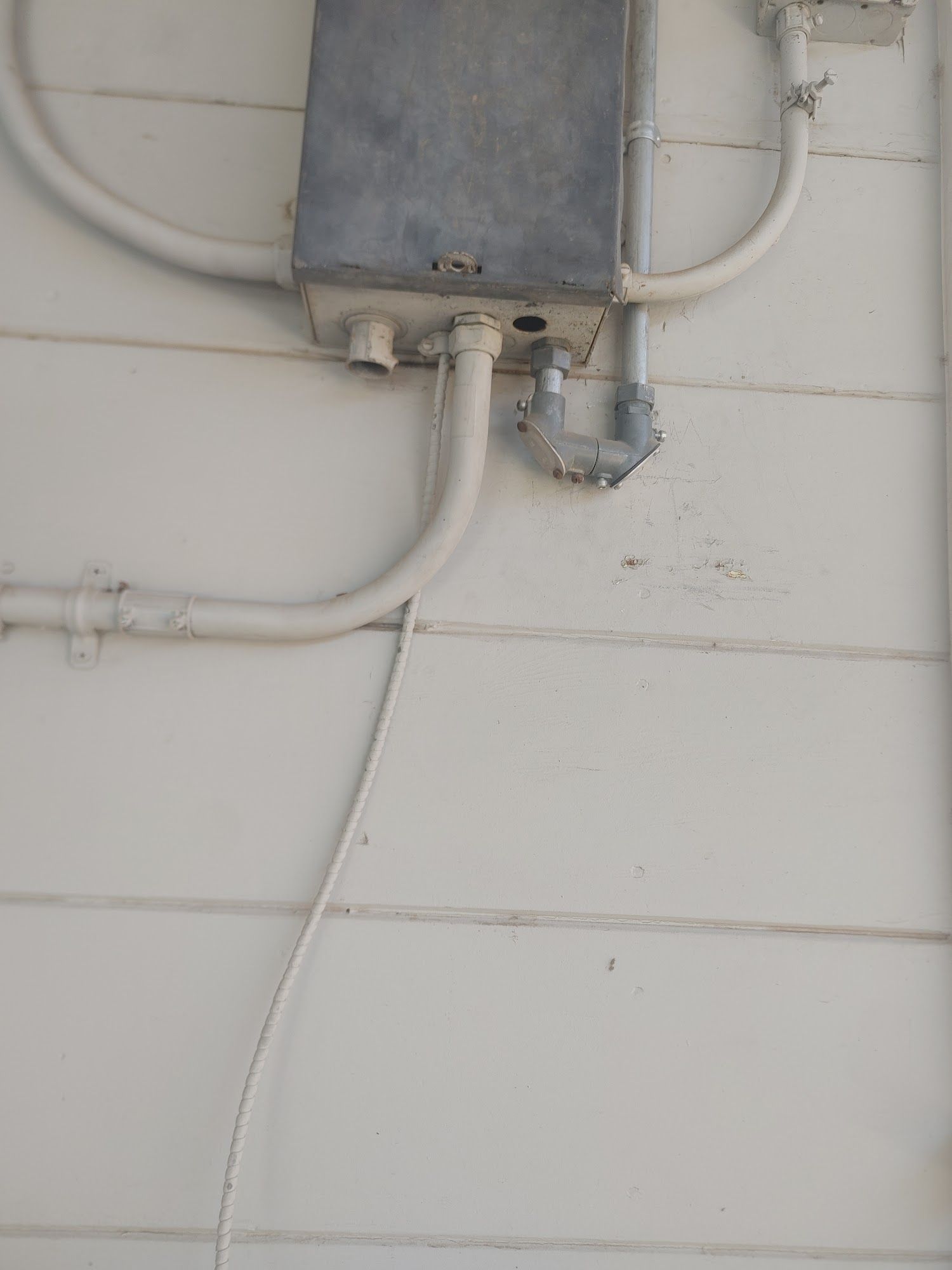 Gray metal electrical box mounted on a white, horizontally-paneled wall with conduit pipes.