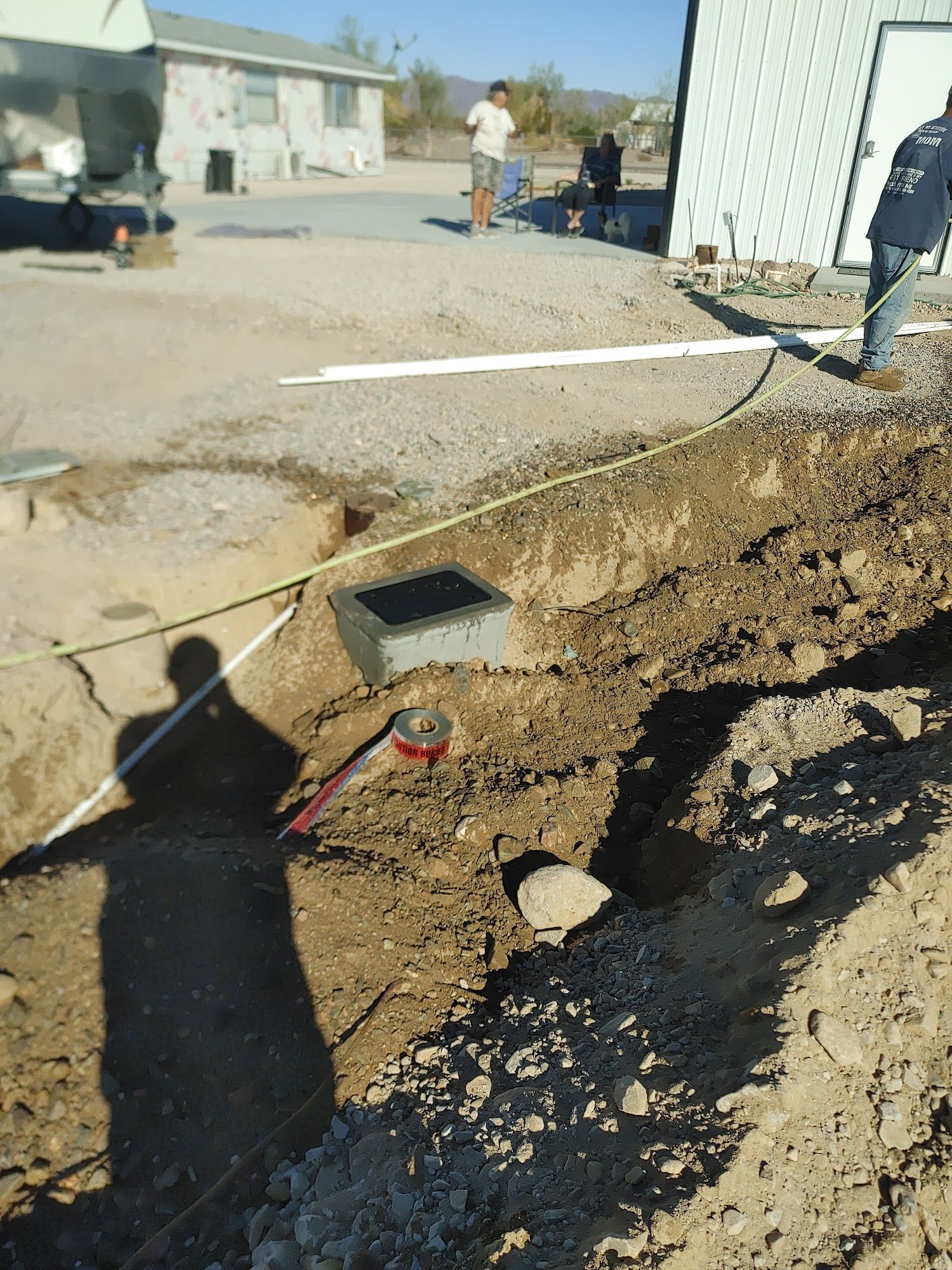Trench with concrete box, pipes, and workers near a building. Outdoor setting, sunny.