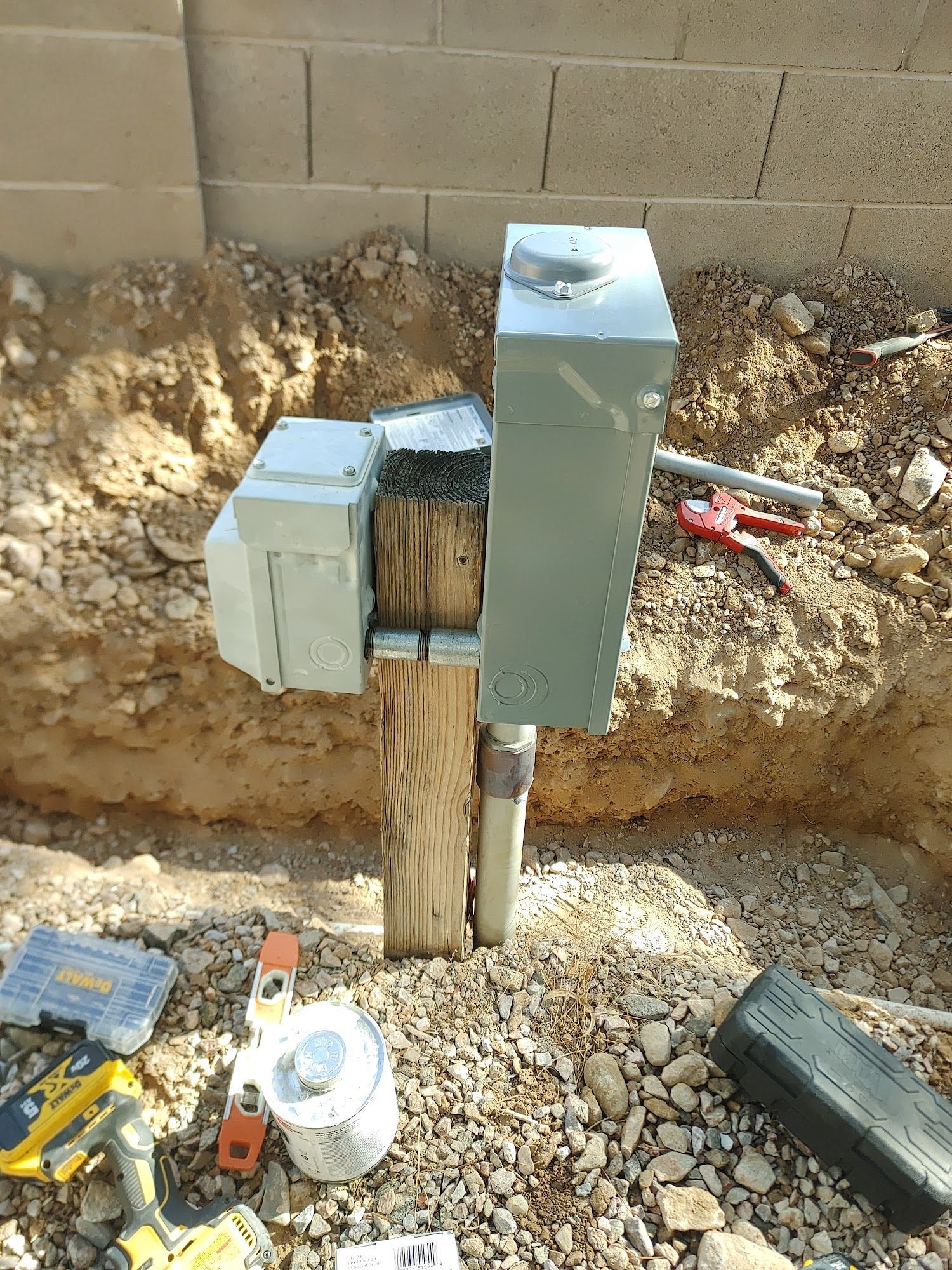 Electrical box attached to a wooden post near a brick wall and dirt ground with tools.