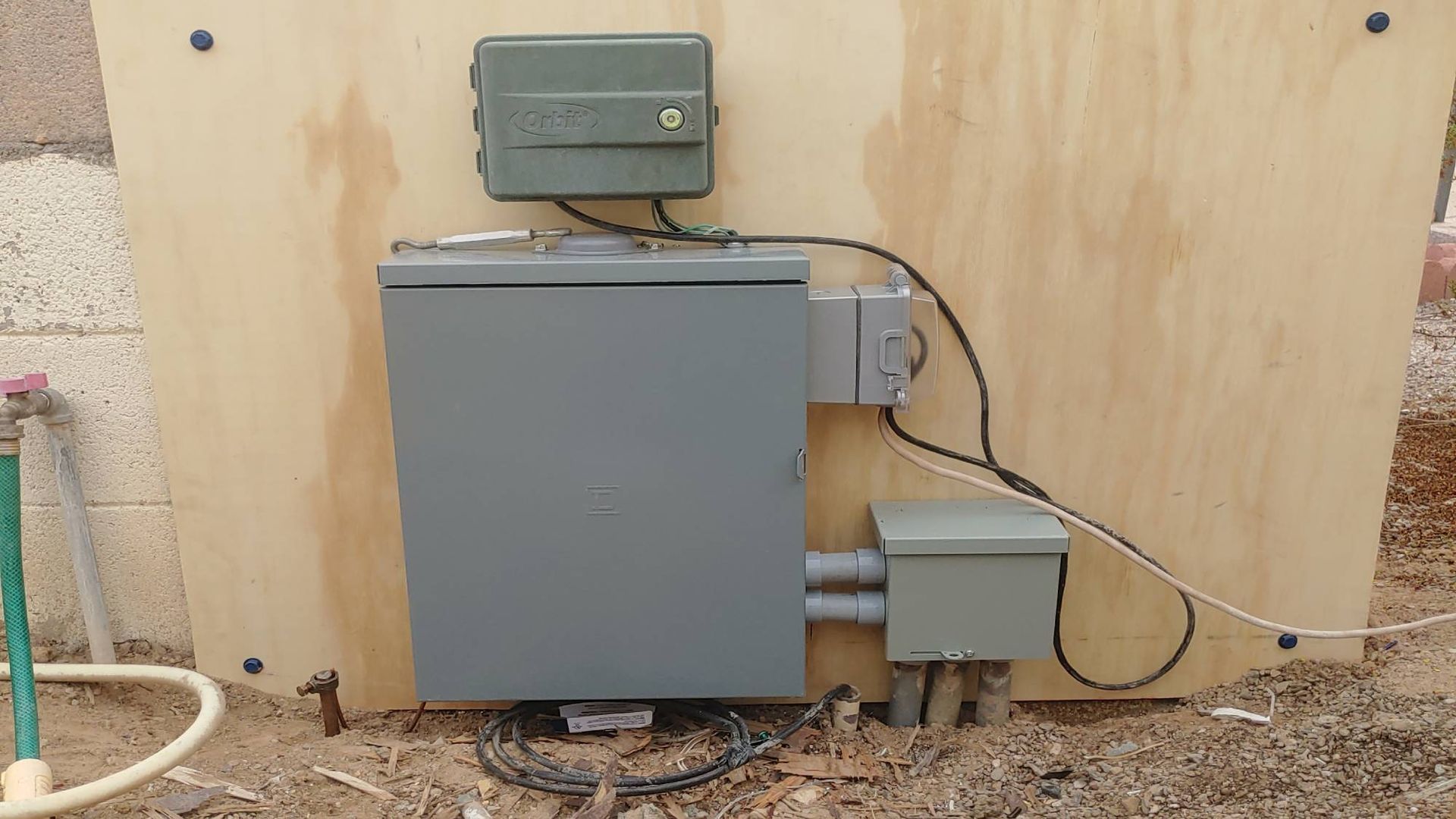 Electrical equipment mounted on a wooden panel: gray boxes and wiring near a brick wall and dirt ground.