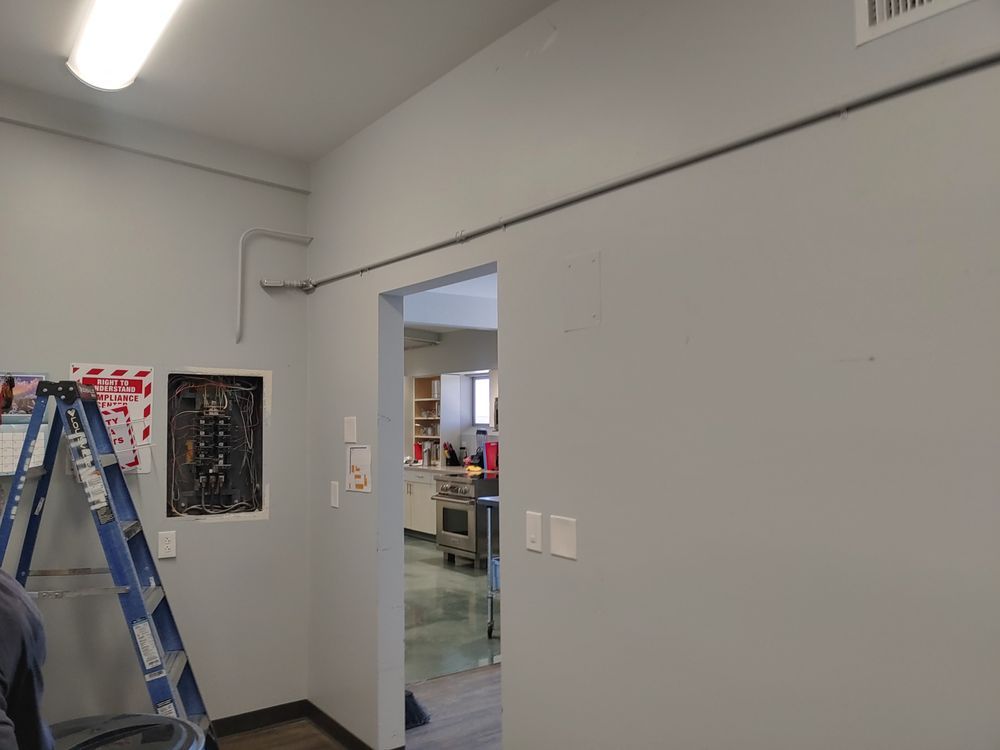Electrical conduit runs across a gray wall, near a doorway and a circuit breaker panel with a ladder.