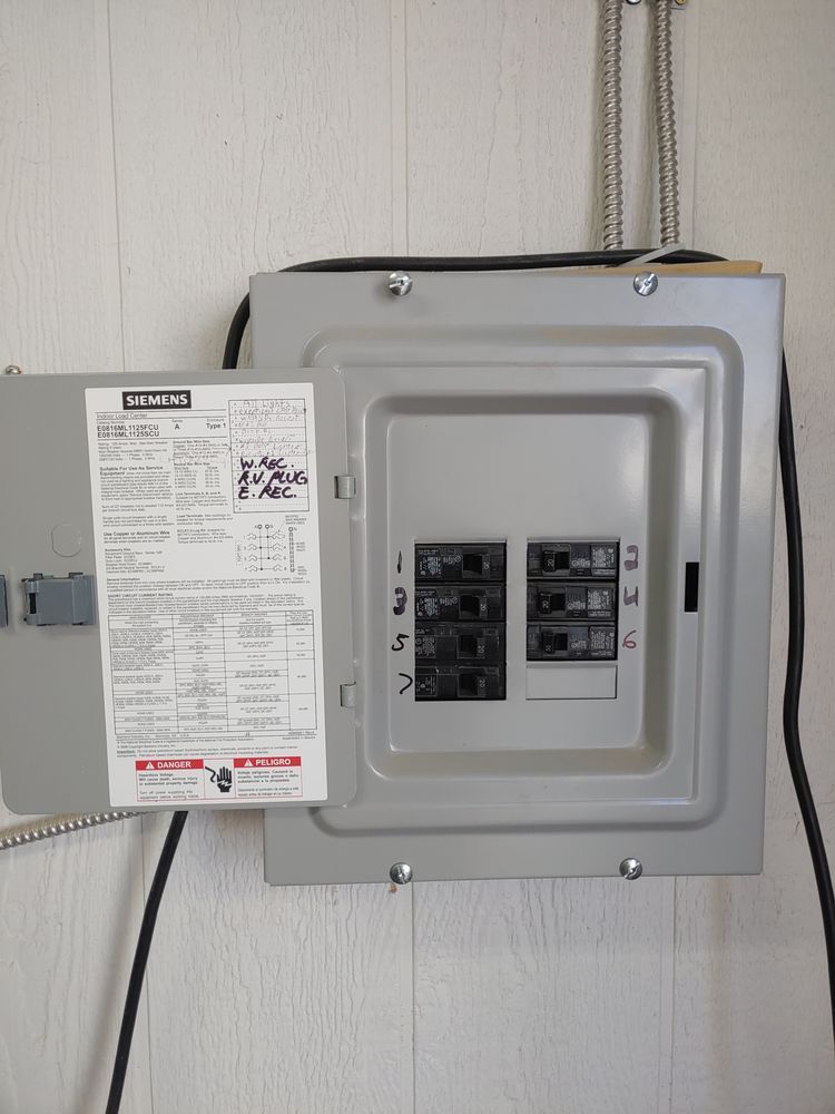 Electrical panel on a white wall. The panel door is open, revealing circuit breakers and a label with text.