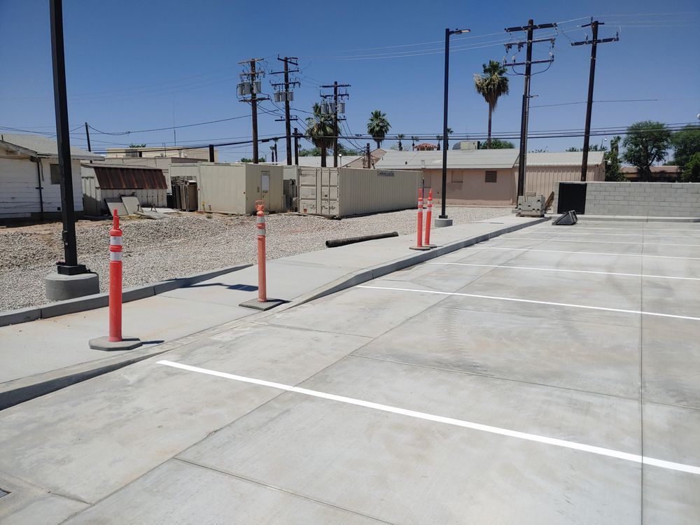 Concrete parking lot with orange traffic cones along a curb. Power lines and buildings in the background.