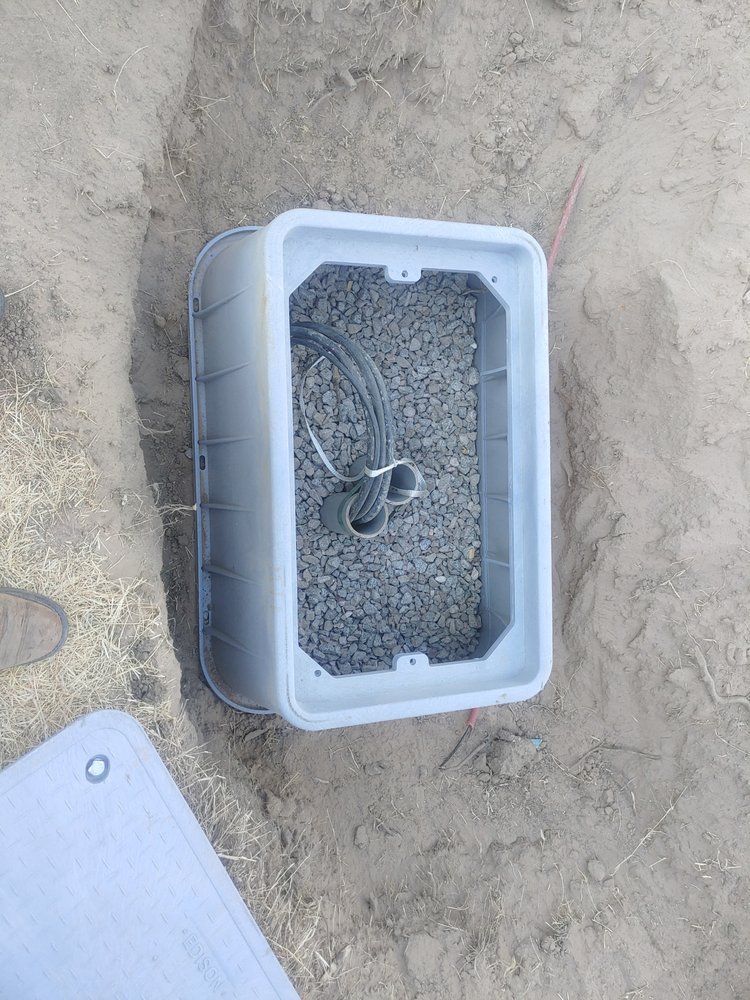 Open gray plastic utility box in a shallow trench; contains cables and gravel.