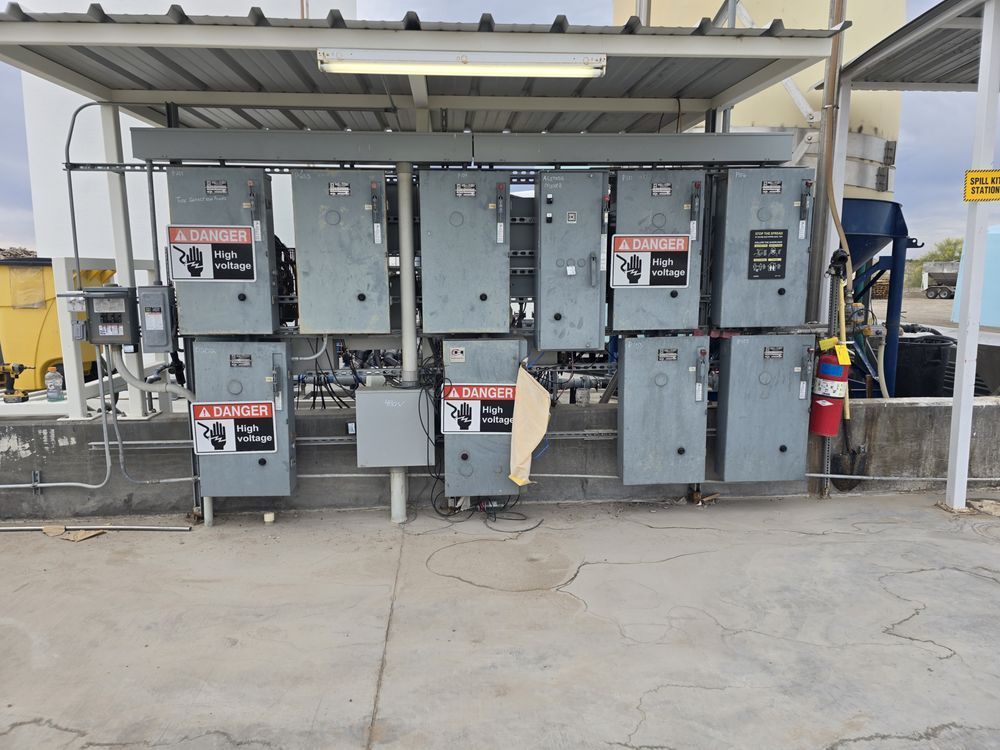 Electrical control boxes mounted outdoors under a metal awning. Concrete base with conduits and wires.