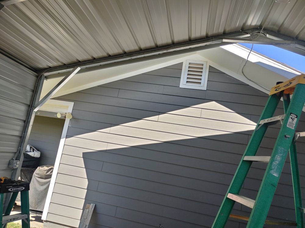 Gray siding and white trim on a building with a metal carport roof; a ladder is positioned nearby.