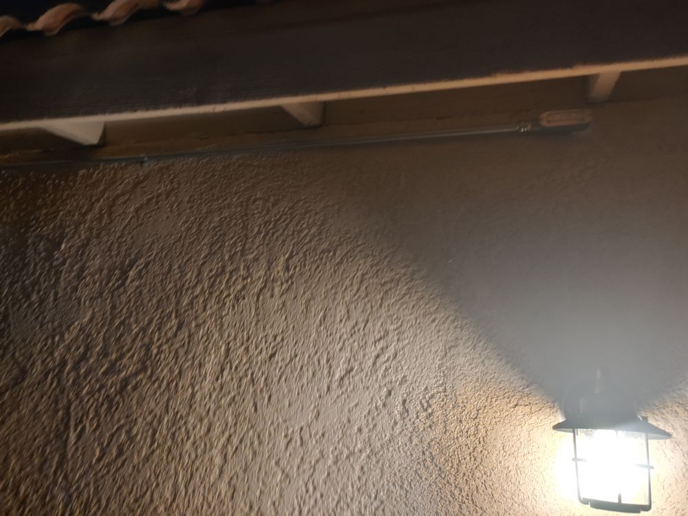 Outdoor wall with textured stucco, lit by a lantern casting a shadow. A wooden beam is above.