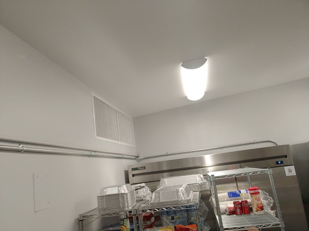 A commercial kitchen corner with a refrigerator, shelves, and a ceiling light.