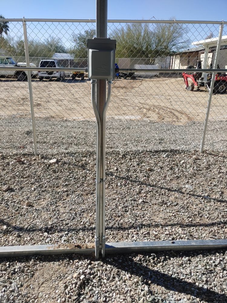 A shiny metal pole with a sensor attached stands in a gravel lot, next to a chain-link fence.