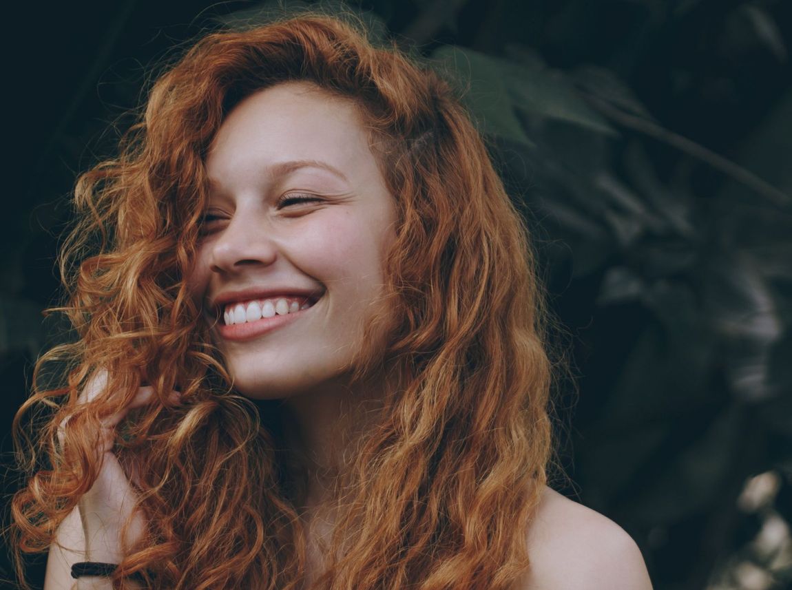 Woman with curly red hair smiles widely, eyes closed, against a blurred green backdrop.