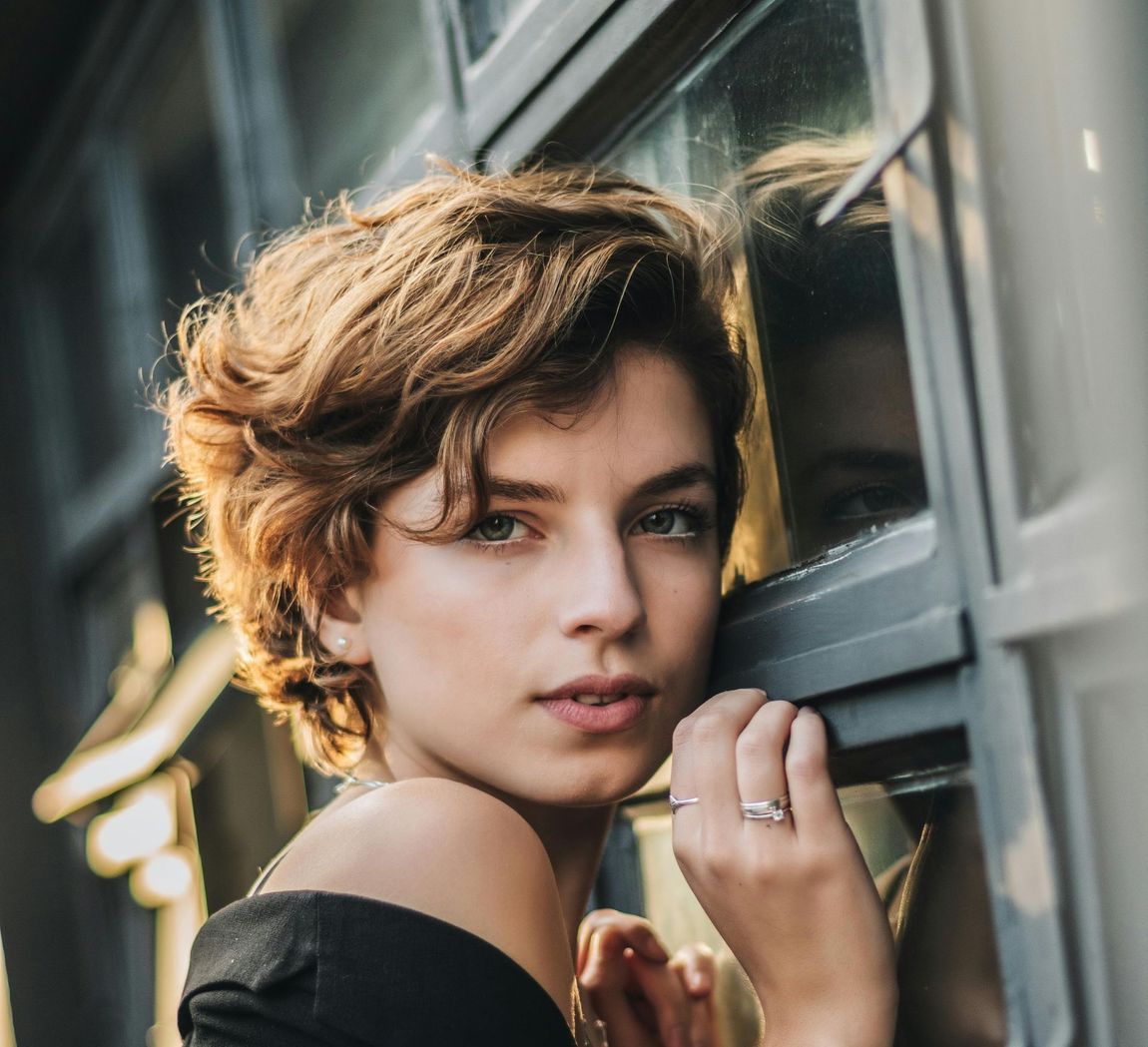 Woman with short brown curly hair, looking toward camera, hand on window frame, sunlight.