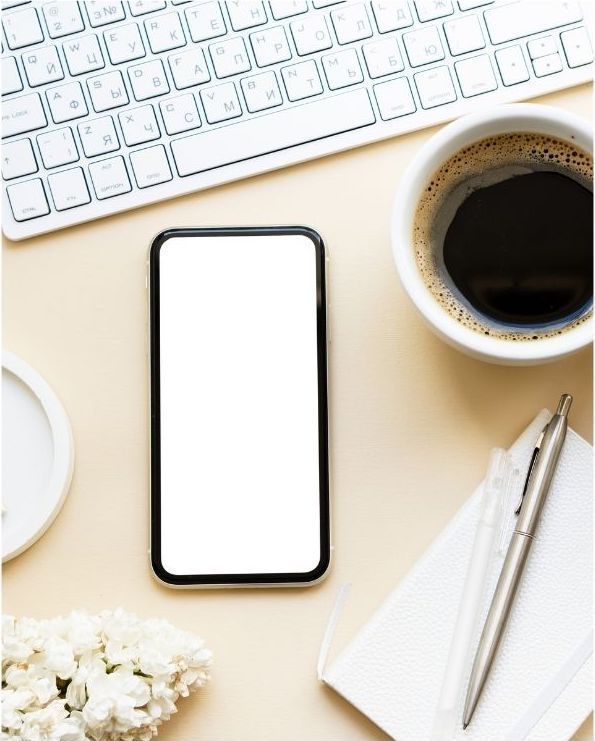 A cell phone with a white screen is sitting on a desk next to a cup of coffee and a keyboard.