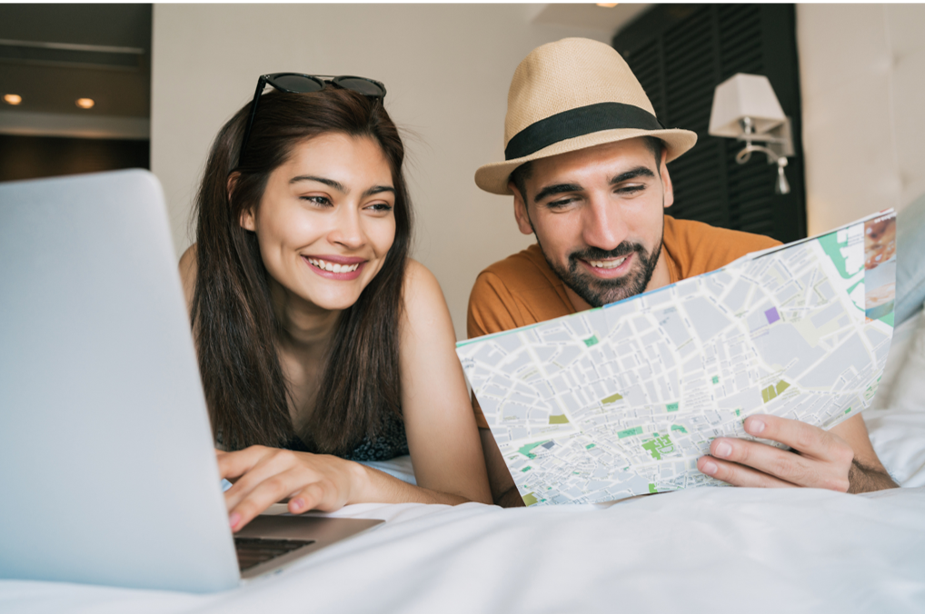 A man and a woman are laying on a bed looking at a map and a laptop.