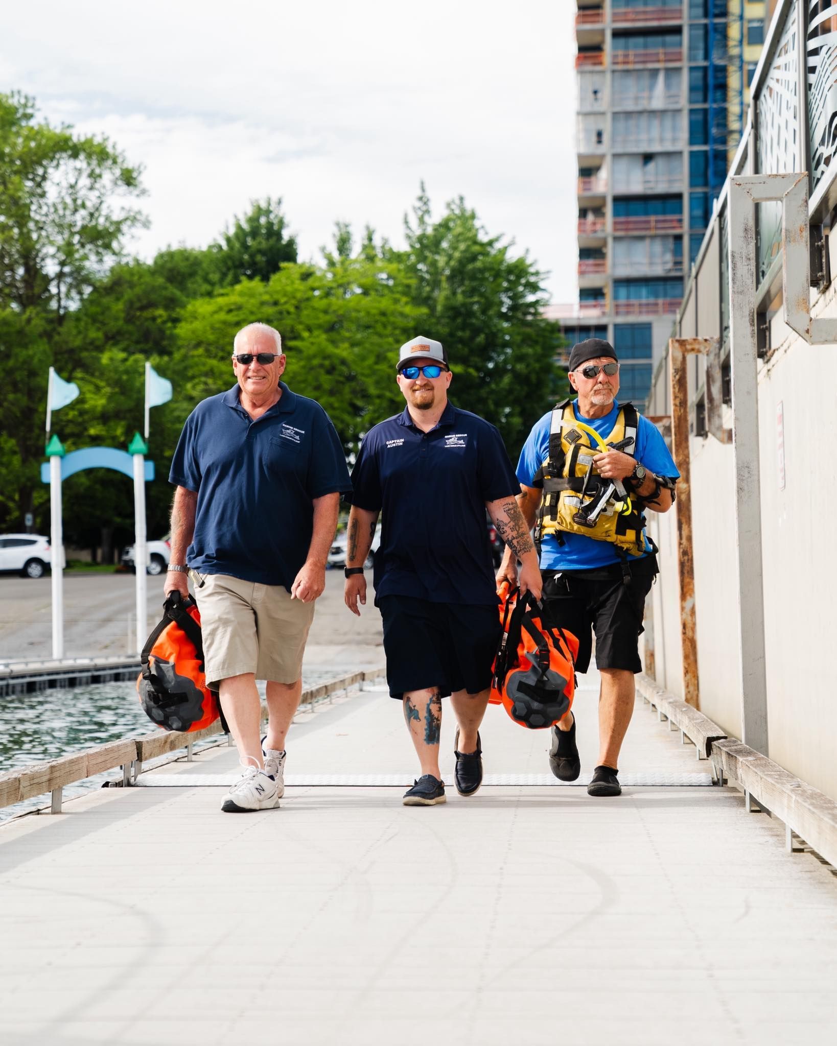 Austin Munda with two  men in blue shirts and shorts walk on a dock, carrying orange bags, near water and a tall building.