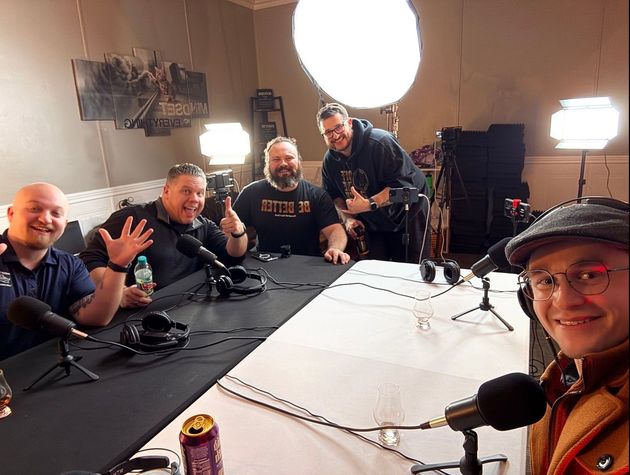 Five men at a podcast recording table with microphones, smiling. Studio setting with lights and equipment.