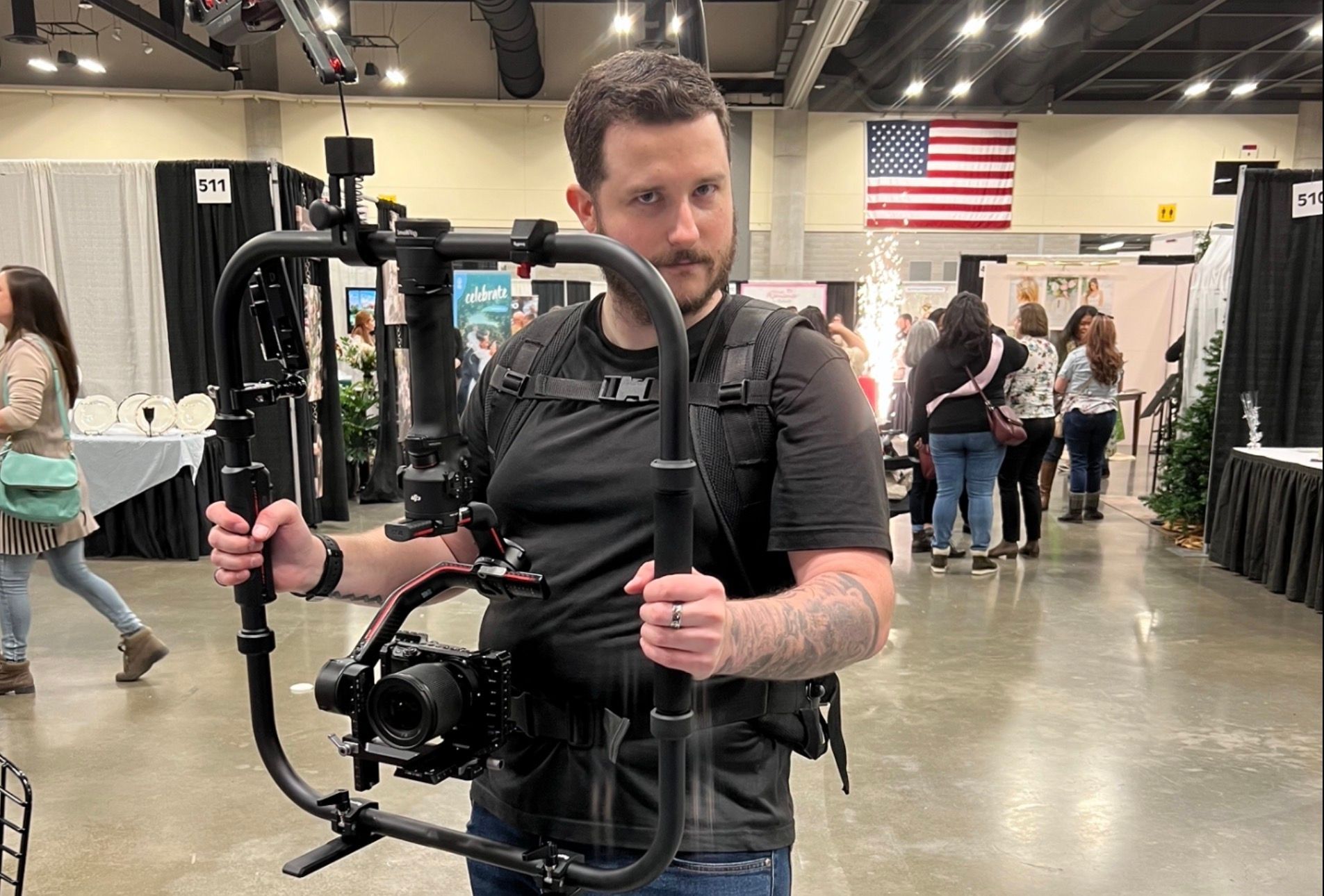 Joshua Miller holding a camera stabilizer, indoors, looking at the camera. An American flag hangs in the background.