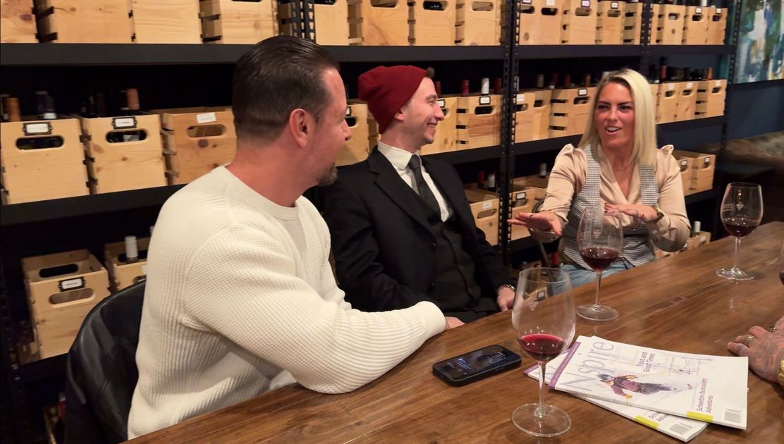 People at bar, two shaking hands. Others seated, some using a laptop, in a dimly lit setting.