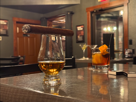Cigar balanced on a whiskey glass, with a cocktail and a lit room in the background.