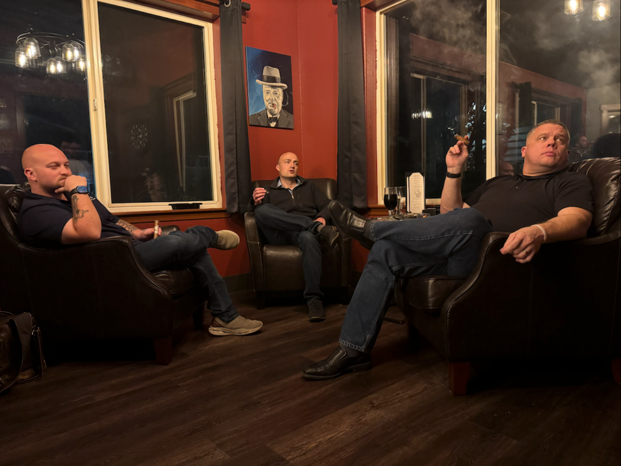 Three men relaxing in leather chairs, smoking cigars in a dimly lit lounge. Red walls, portrait, window.