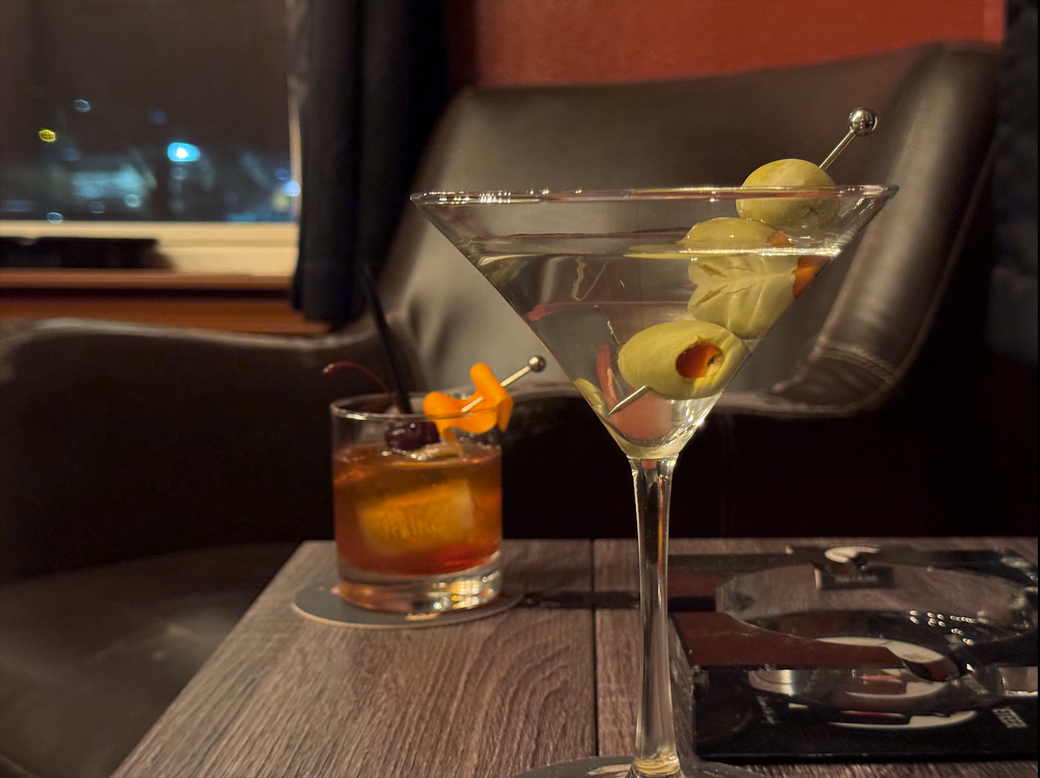 Martini with olives and Old Fashioned on a table in a dimly lit bar setting.