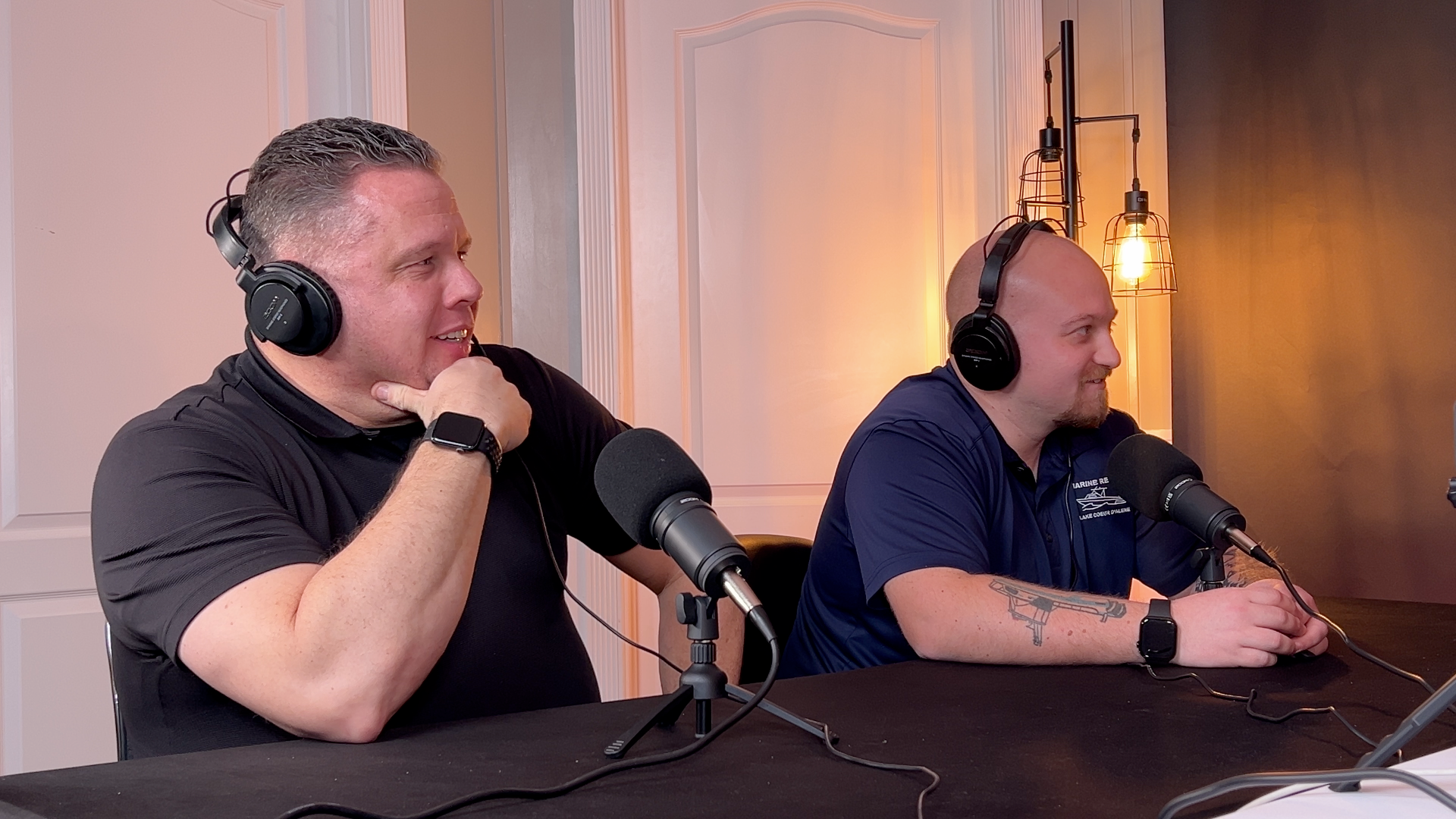 Two men in a studio wearing headphones, speaking into microphones. One rests his chin on his hand.