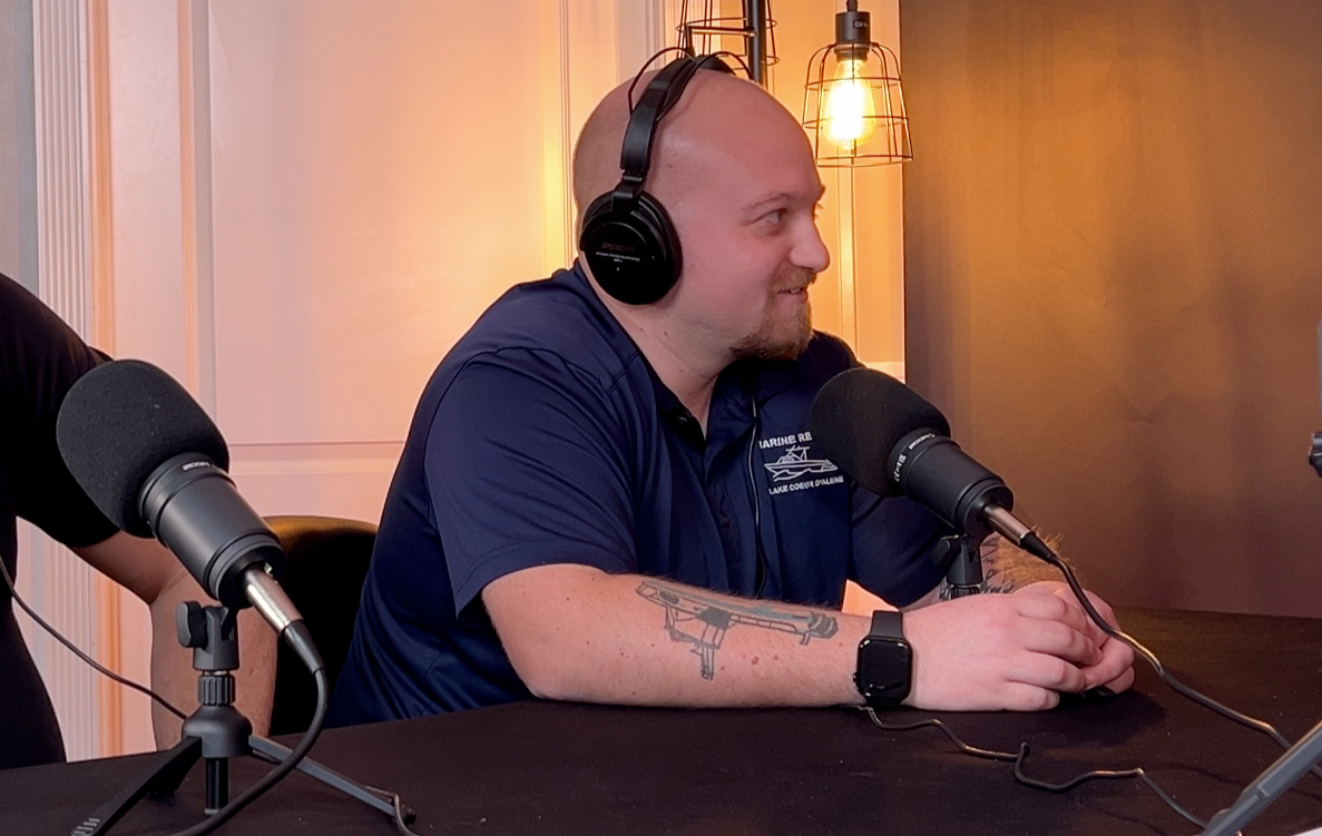 Austin Munda with headphones at a podcast table, speaking into a microphone. Dark blue shirt, tattoo, watch, microphones.