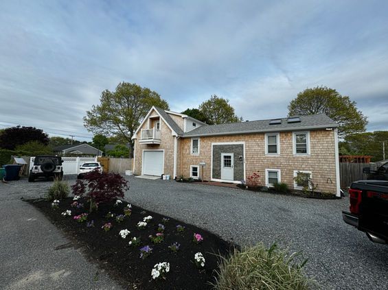 A light brown shingled building with a driveway, flower bed, and parked cars under a cloudy sky.