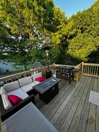 Wooden deck with outdoor seating, fire pit, grill, and heater, surrounded by trees.