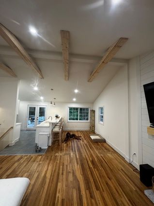 Open-concept living space with exposed wooden beams, hardwood floors, a white kitchen, and a dog lying on the floor.