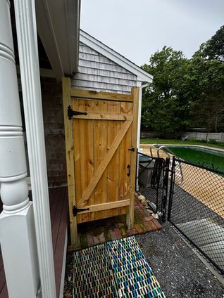 Wooden outdoor shower next to a house with a brick path and fence.