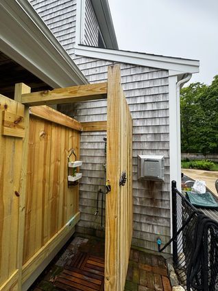 Wooden outdoor shower next to a shingled building, door open.