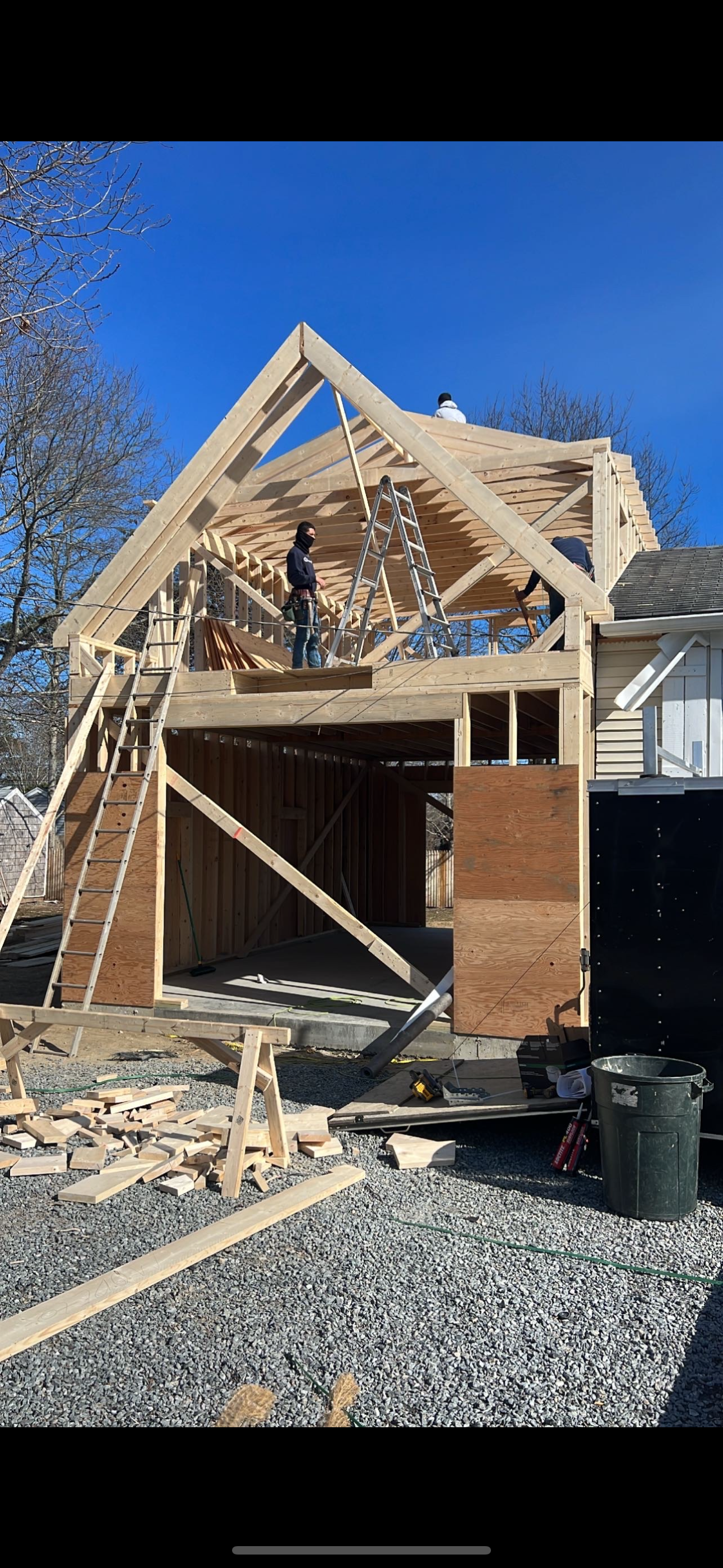 Construction of a wood-framed building. A worker stands on the roof frame. Plywood and gravel at the base. Blue sky.