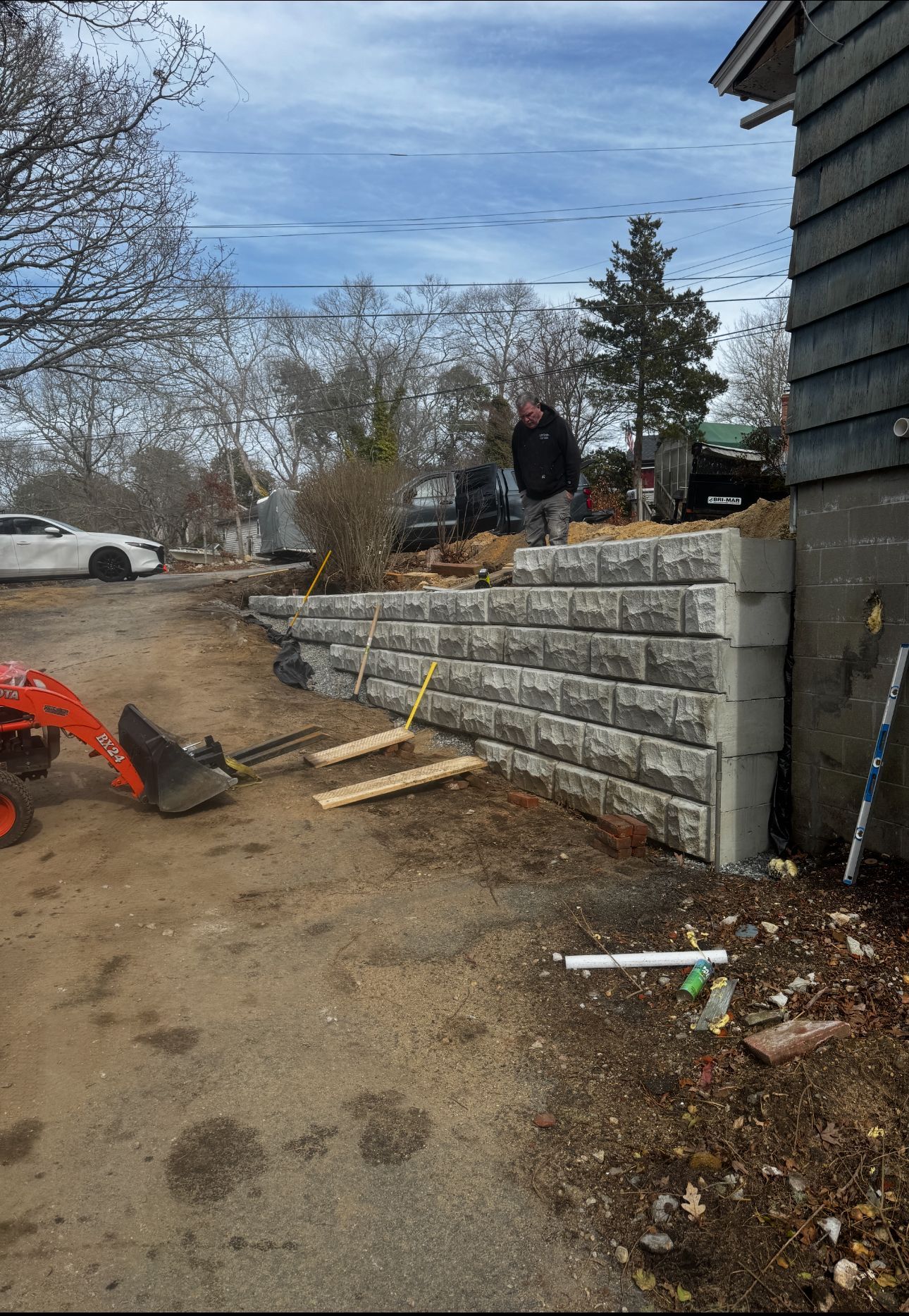 A retaining wall made of gray blocks is being built next to a house. A person stands on top.