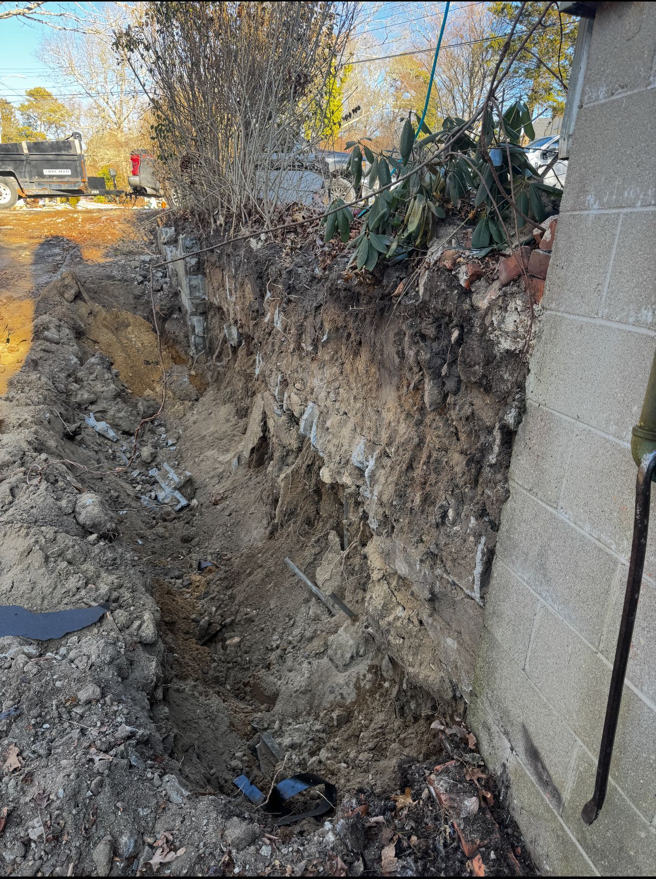 Trench dug next to a concrete wall. Earth, roots, and debris visible. Overcast day.