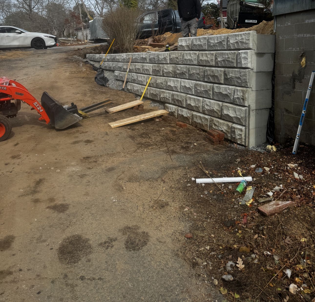 Retaining wall construction: Gray blocks, dirt road, small tractor, building in the background.