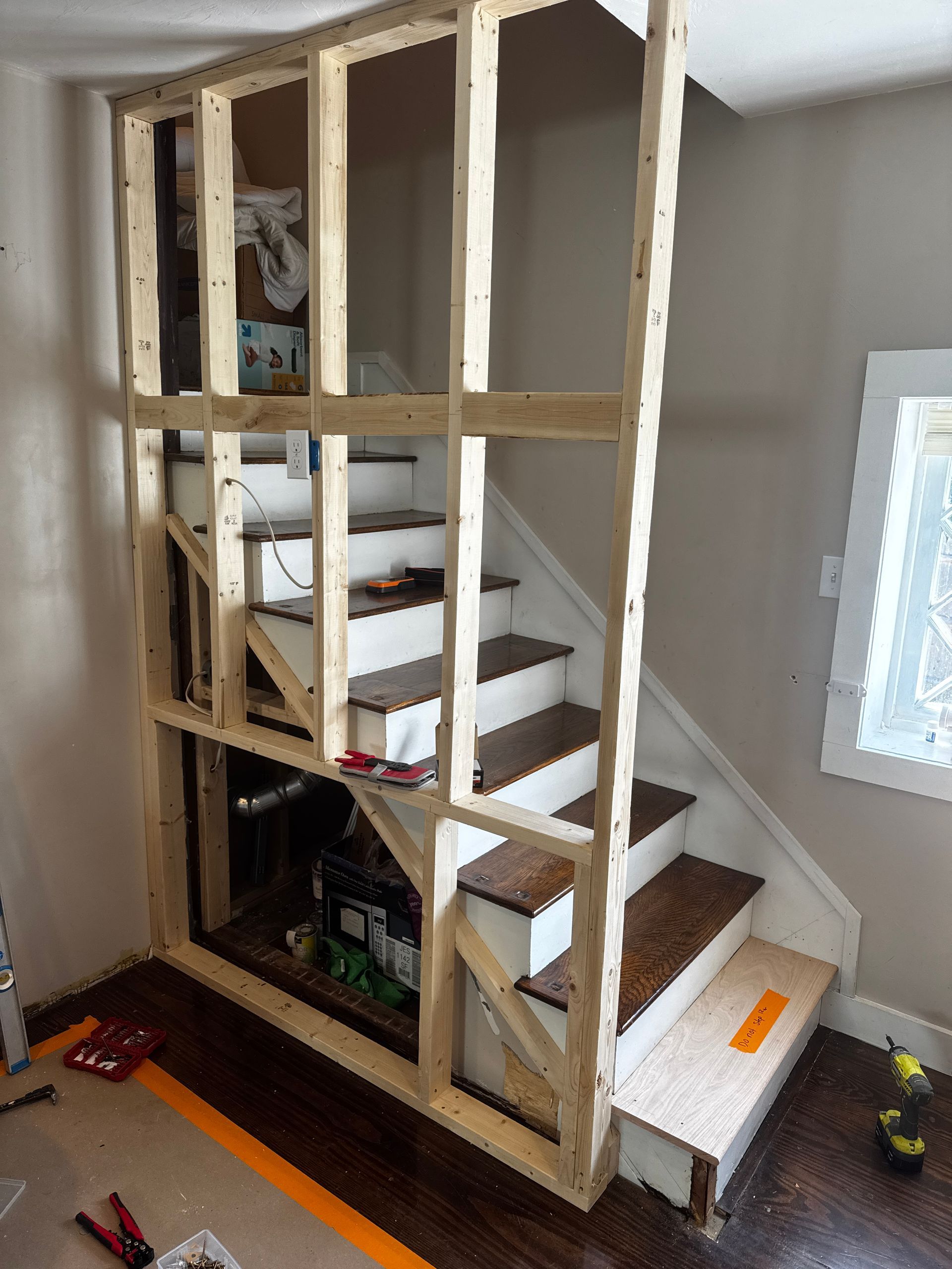 Wooden frame built around a staircase; construction site. Staircase with white steps, dark brown treads, and a window in the background.
