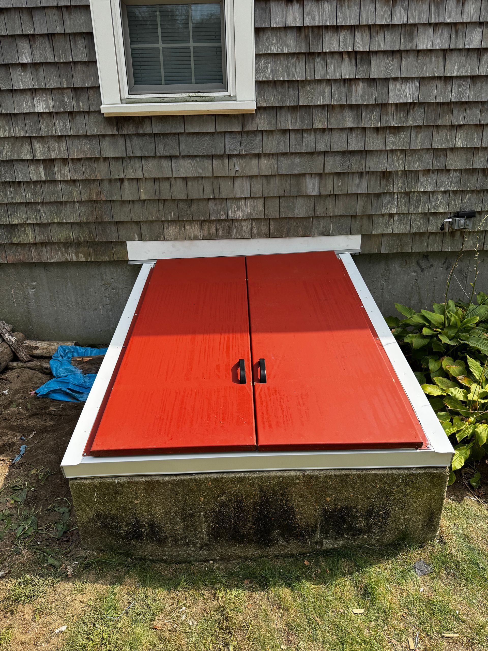 Red double doors on a concrete basement entrance, beneath a weathered wood-shingled wall.