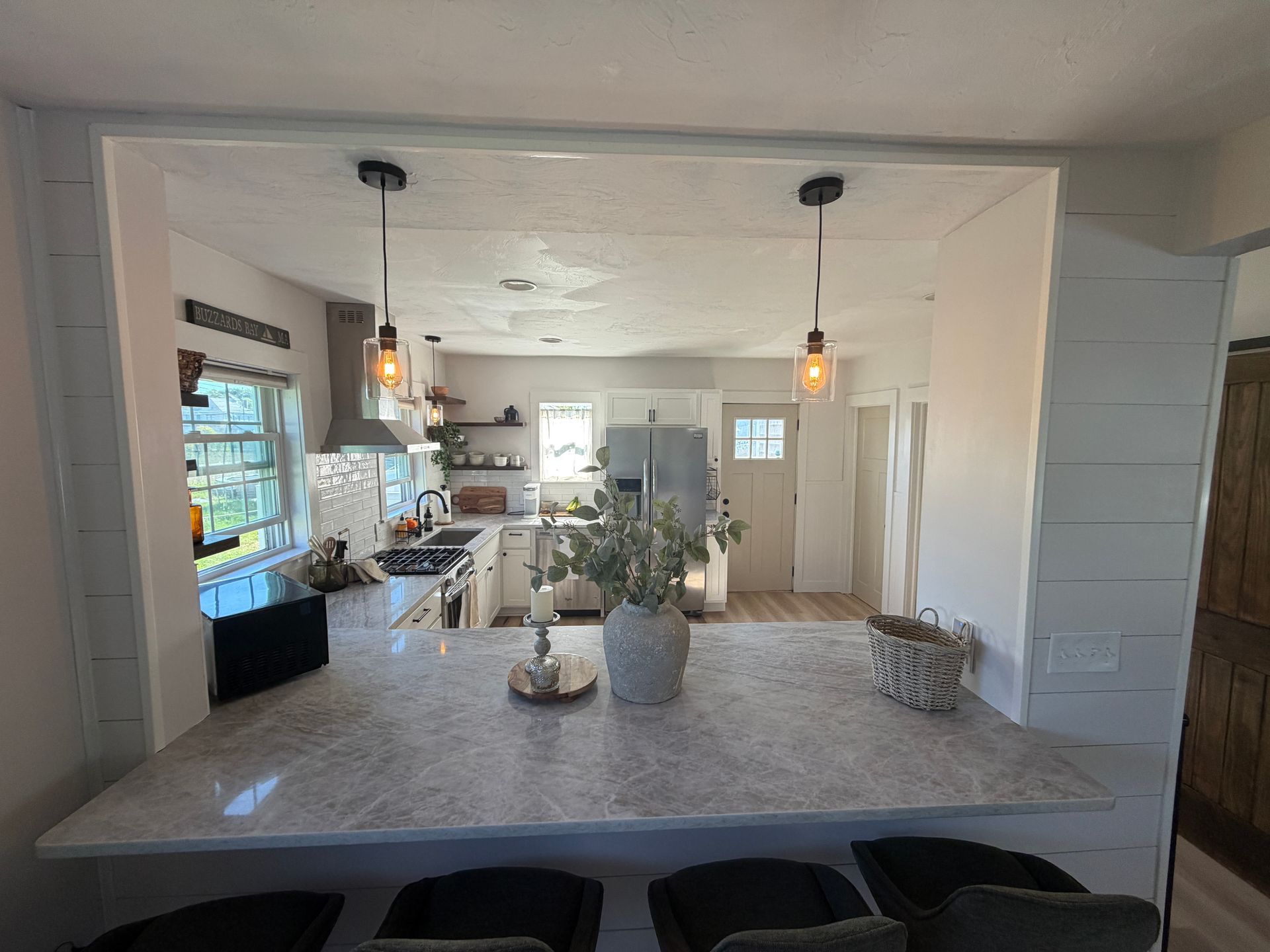 Kitchen with a stone countertop, bar stools, and pendant lights overlooking a bright, white kitchen.