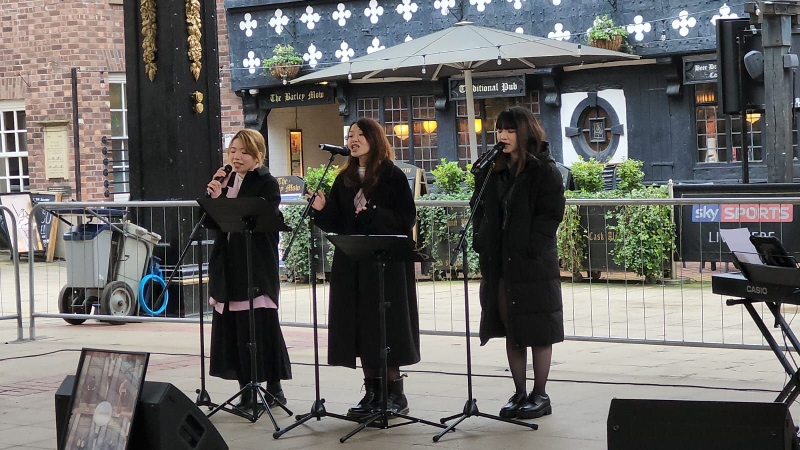 Three women singing on a stage in front of a building. They wear dark coats and use microphones.