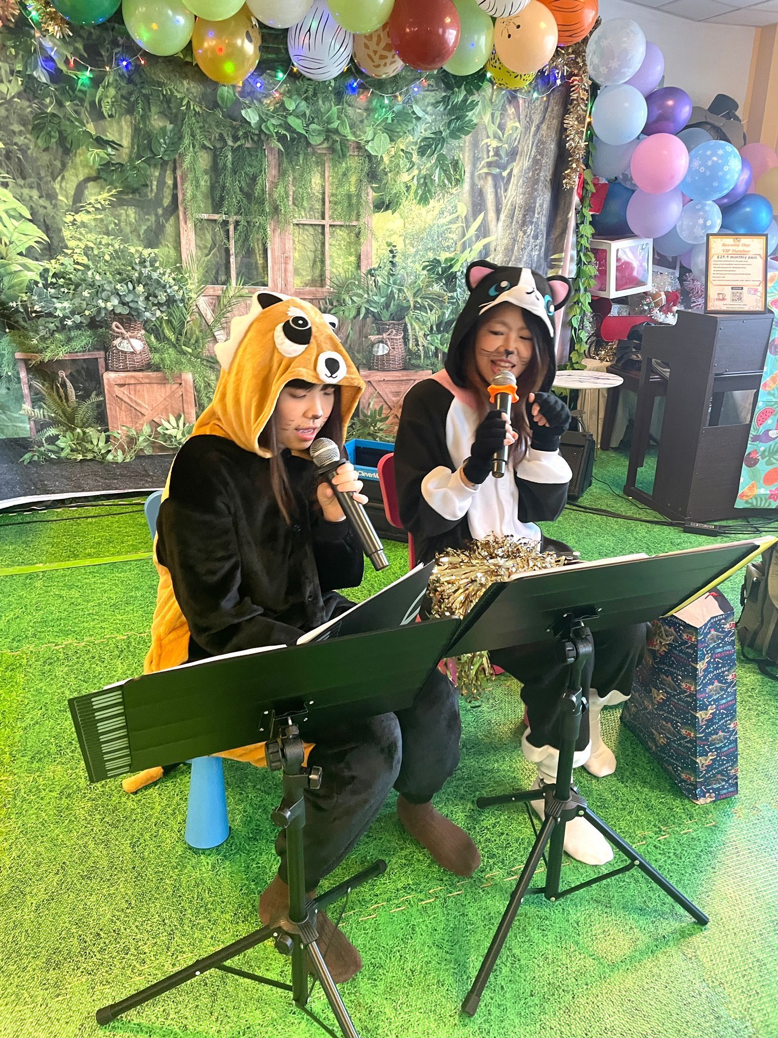 Two people in costumes singing with microphones at a green-themed event, with balloons and a backdrop of a garden.