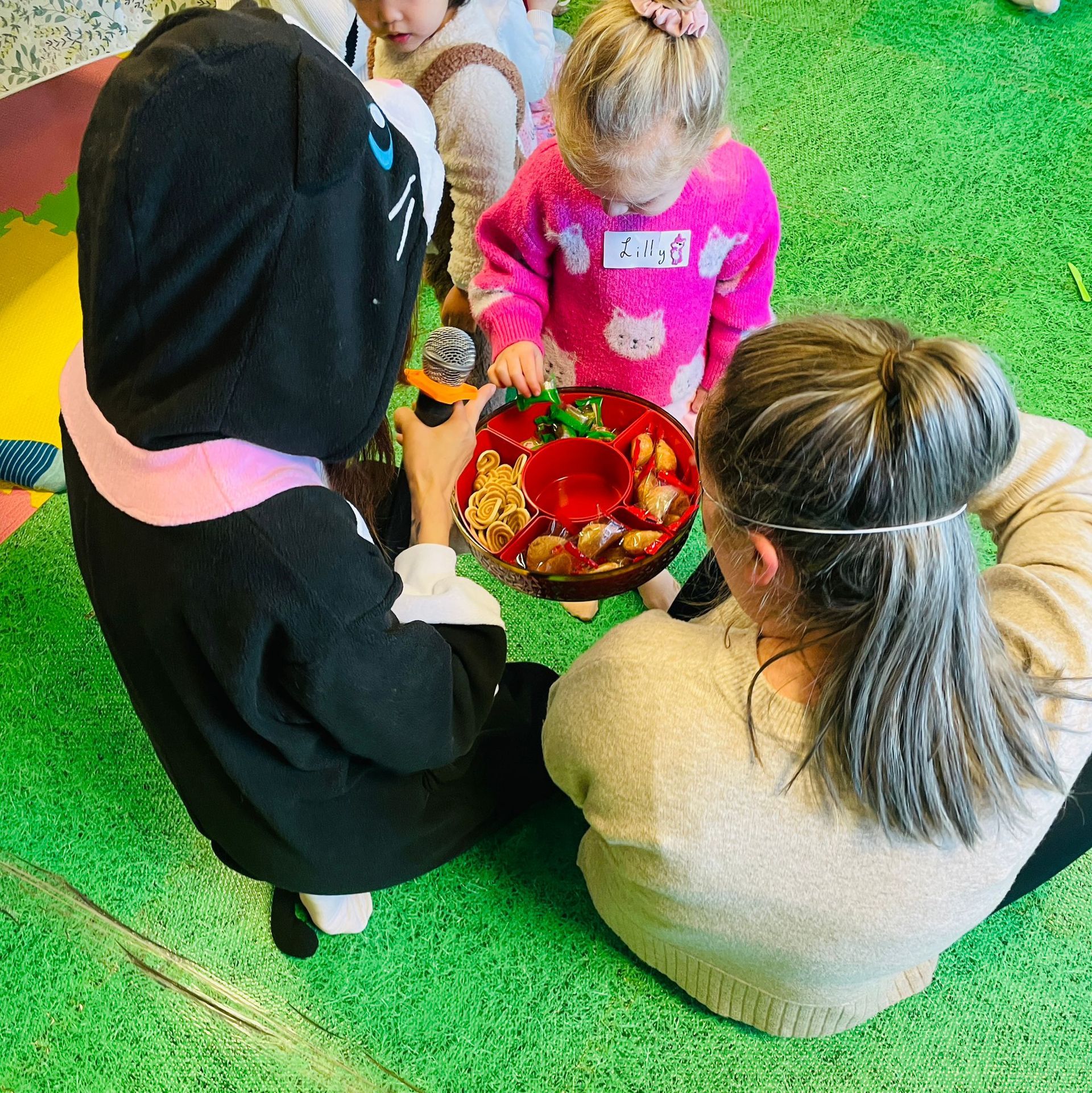 Children and adult gather around a tray of food on green grass. One child in a cat costume, another in pink sweater.